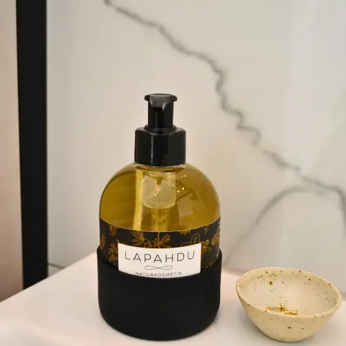 LAPAHDU Flüssigseife 250 ml in Glasflasche mit schwarzer Silikonhülle am Waschbecken, daneben ein kleines Schälchen mit Schmuck